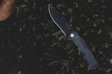 A black folding tactical knife is placed on dark foliage with a textured, serrated black handle and a shiny, curved blade. The background includes green leaves, contributing to a rugged, outdoor setting.