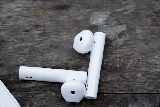 Wireless earbuds are placed on a textured wooden surface. The earbuds are white with metallic and black accents on them, creating a contrast with the rustic background.