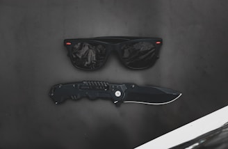 A pair of black sunglasses with reflective lenses placed above a black folding knife on a matte dark surface. Tiny red accents appear on the corners of the sunglasses.