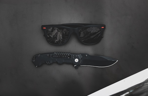 A pair of black sunglasses with reflective lenses placed above a black folding knife on a matte dark surface. Tiny red accents appear on the corners of the sunglasses.