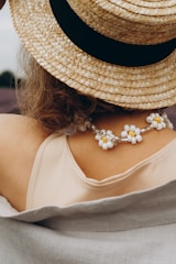 A person is wearing a straw hat with a wide black band, a cream-colored top, and floral bead jewelry draped around their neck. Part of a blanket or fabric covers their shoulders.