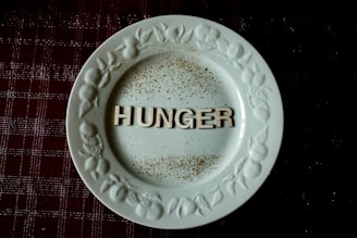 A white plate with ornate designs along the rim features the word 'HUNGER' in bold letters at the center. The plate is placed on a dark, textured surface, possibly a tablecloth, with scattered specks resembling spices or dirt around the letters.