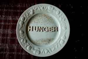 A white plate with ornate designs along the rim features the word 'HUNGER' in bold letters at the center. The plate is placed on a dark, textured surface, possibly a tablecloth, with scattered specks resembling spices or dirt around the letters.