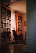 A cozy reading nook featuring bookshelves filled with numerous books. A soft light filters through a sheer curtain next to a window, illuminating a red armchair on a rug. The space conveys warmth and comfort.