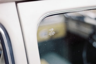 The image depicts a close-up view of a vintage car window, displaying its reflective glass surface. The interior and exterior details of the vehicle are blurred, creating a soft focus on the etched writing in the window that includes a logo and specifications.