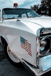 A vintage white police car is parked outdoors, featuring a retro design with a chrome finish. The front bumper and headlight are prominently visible, along with a decal of an American flag on the side. The car is equipped with a red emergency light on top, and the scene is surrounded by trees under a cloudy sky.