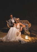 A couple sits closely on a wicker bench at night. The woman is wearing a flowing white dress, and the man is in a gray suit. A decorative lantern with a lit candle is placed on the ground beside them, casting a warm glow that highlights their faces and attire.