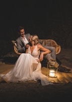 A couple sits closely on a wicker bench at night. The woman is wearing a flowing white dress, and the man is in a gray suit. A decorative lantern with a lit candle is placed on the ground beside them, casting a warm glow that highlights their faces and attire.