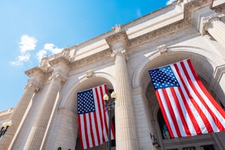Two large American flags hang prominently on a grand, classical-style building with tall columns and intricate architectural details. The sky is clear and blue, adding to the sense of openness and grandeur. The scene conveys a sense of patriotism and stateliness.