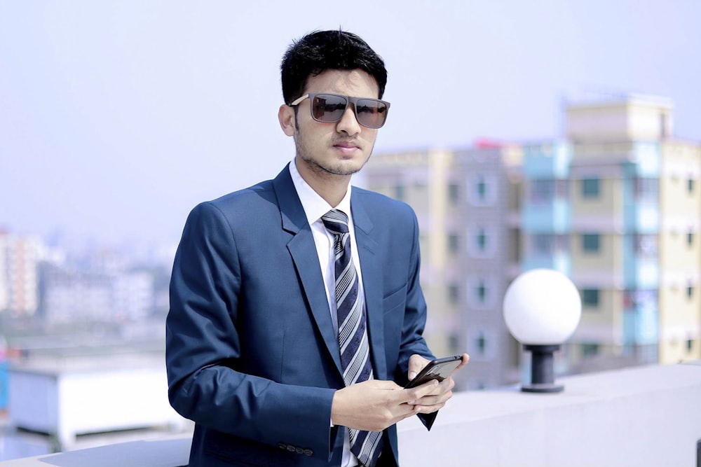 man in blue suit jacket wearing black sunglasses holding black smartphone