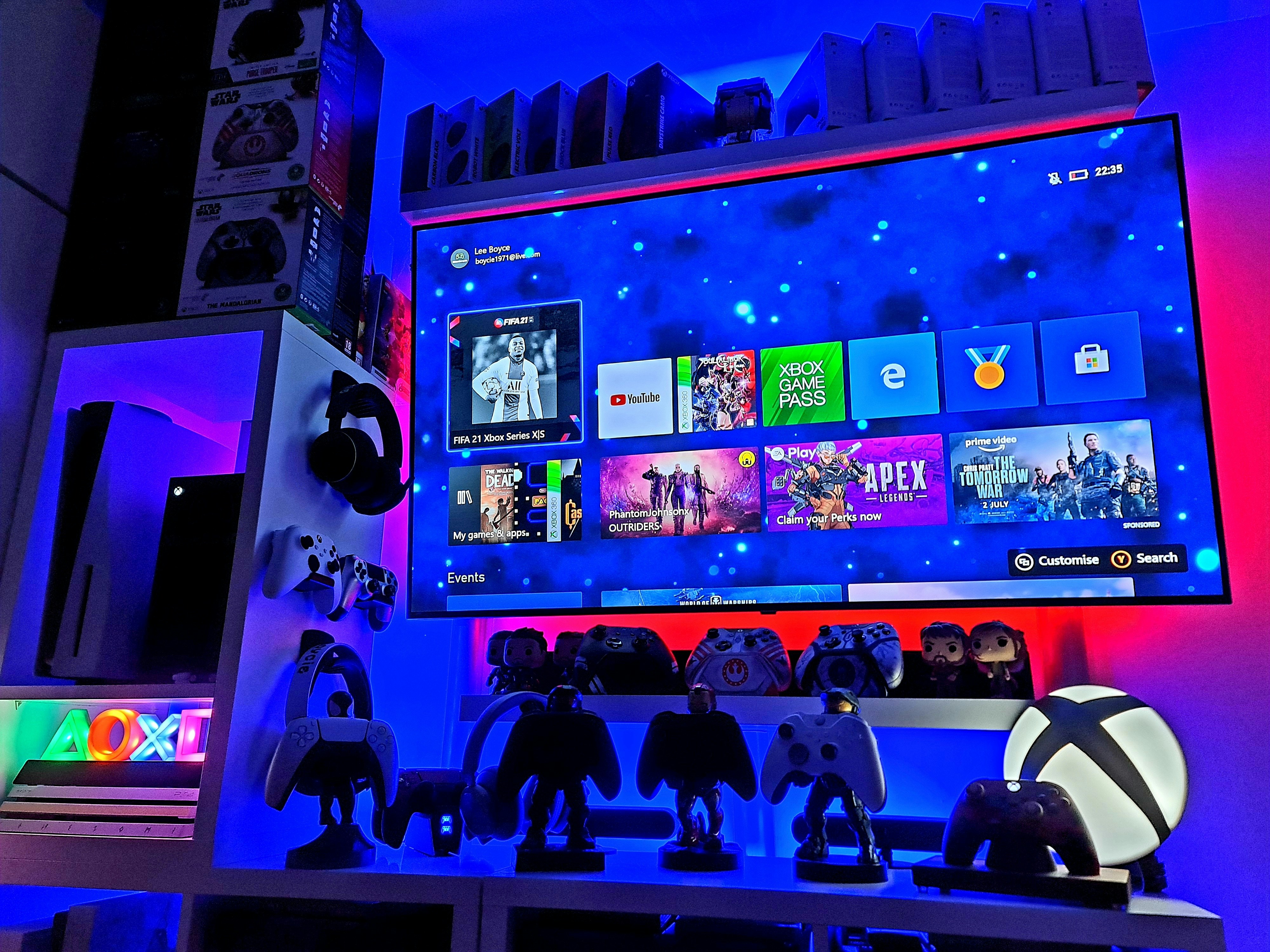 A modern gaming setup featuring a large wall-mounted screen displaying a gaming console interface. The room is decorated with gaming-themed items such as controllers and collectibles. The ambient lighting includes shades of blue and red, creating a vibrant atmosphere.