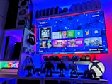 A modern gaming setup featuring a large wall-mounted screen displaying a gaming console interface. The room is decorated with gaming-themed items such as controllers and collectibles. The ambient lighting includes shades of blue and red, creating a vibrant atmosphere.