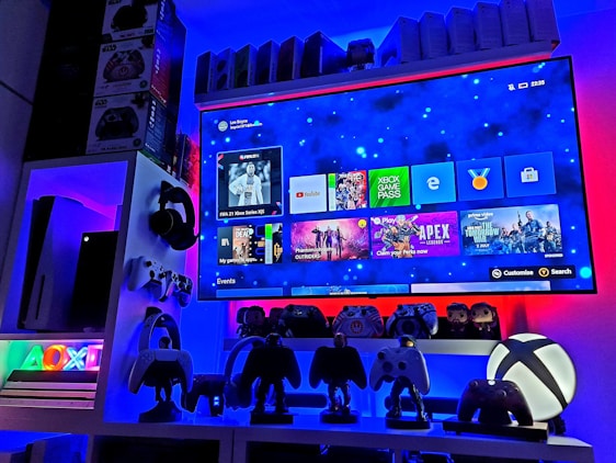 A modern gaming setup featuring a large wall-mounted screen displaying a gaming console interface. The room is decorated with gaming-themed items such as controllers and collectibles. The ambient lighting includes shades of blue and red, creating a vibrant atmosphere.