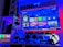 A modern gaming setup featuring a large wall-mounted screen displaying a gaming console interface. The room is decorated with gaming-themed items such as controllers and collectibles. The ambient lighting includes shades of blue and red, creating a vibrant atmosphere.