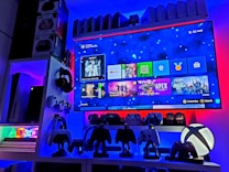A modern gaming setup featuring a large wall-mounted screen displaying a gaming console interface. The room is decorated with gaming-themed items such as controllers and collectibles. The ambient lighting includes shades of blue and red, creating a vibrant atmosphere.