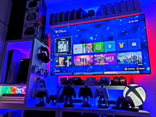 A modern gaming setup featuring a large wall-mounted screen displaying a gaming console interface. The room is decorated with gaming-themed items such as controllers and collectibles. The ambient lighting includes shades of blue and red, creating a vibrant atmosphere.