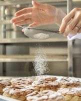 A pair of hands sifts powdered sugar onto pastries arranged neatly on a tray in a kitchen setting. The lightly powdered pastries seem freshly baked and inviting.