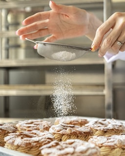 A pair of hands sifts powdered sugar onto pastries arranged neatly on a tray in a kitchen setting. The lightly powdered pastries seem freshly baked and inviting.