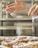 A pair of hands sifts powdered sugar onto pastries arranged neatly on a tray in a kitchen setting. The lightly powdered pastries seem freshly baked and inviting.