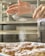 A pair of hands sifts powdered sugar onto pastries arranged neatly on a tray in a kitchen setting. The lightly powdered pastries seem freshly baked and inviting.