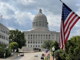 A large domed government building with classical architecture is visible in the background. An American flag prominently waves in the foreground on the right. The scene includes trees, a few people walking on the sidewalk, and cars parked along the street leading to the building.