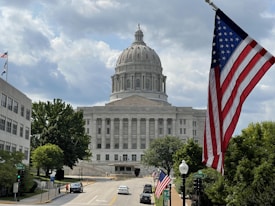 A large domed government building with classical architecture is visible in the background. An American flag prominently waves in the foreground on the right. The scene includes trees, a few people walking on the sidewalk, and cars parked along the street leading to the building.