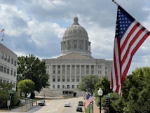 A large domed government building with classical architecture is visible in the background. An American flag prominently waves in the foreground on the right. The scene includes trees, a few people walking on the sidewalk, and cars parked along the street leading to the building.