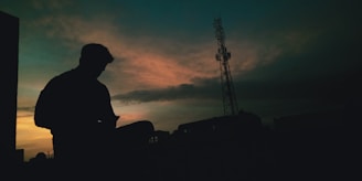 A silhouette of a person sitting while using a mobile phone against a dramatic sunset sky. The sky features shades of orange and blue, with whispy clouds creating a textured backdrop. A tall communication tower stands prominently on the right side of the image, adding an industrial element to the otherwise serene scene.