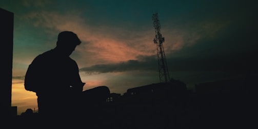 A silhouette of a person sitting while using a mobile phone against a dramatic sunset sky. The sky features shades of orange and blue, with whispy clouds creating a textured backdrop. A tall communication tower stands prominently on the right side of the image, adding an industrial element to the otherwise serene scene.