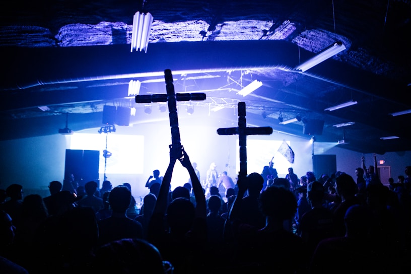 A large crowd gathered in a dark indoor venue, with blue and white lights illuminating the space. Two individuals hold up wooden crosses, and the atmosphere suggests a concert or event taking place. Silhouettes of people are visible against the bright lighting on stage.