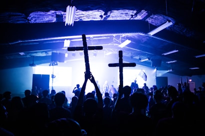 A large crowd gathered in a dark indoor venue, with blue and white lights illuminating the space. Two individuals hold up wooden crosses, and the atmosphere suggests a concert or event taking place. Silhouettes of people are visible against the bright lighting on stage.