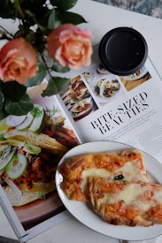 A close-up shot of a magazine spread featuring images of food, accompanied by an article titled 'Bite-Sized Beauties.' A plate with baked bread or pastry with cheese and toppings is positioned over part of the magazine. Beside the plate, there is a black coffee cup lid and a bouquet of peach roses.