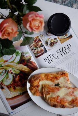 A close-up shot of a magazine spread featuring images of food, accompanied by an article titled 'Bite-Sized Beauties.' A plate with baked bread or pastry with cheese and toppings is positioned over part of the magazine. Beside the plate, there is a black coffee cup lid and a bouquet of peach roses.