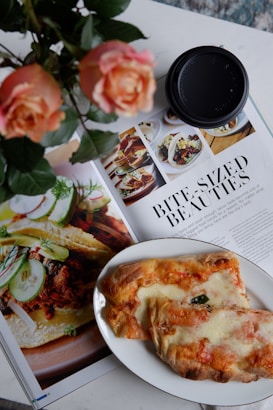 A close-up shot of a magazine spread featuring images of food, accompanied by an article titled 'Bite-Sized Beauties.' A plate with baked bread or pastry with cheese and toppings is positioned over part of the magazine. Beside the plate, there is a black coffee cup lid and a bouquet of peach roses.