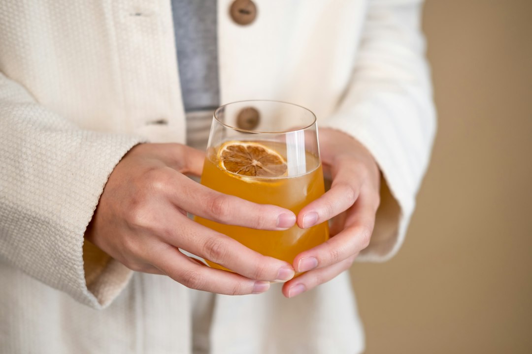 person holding clear drinking glass with orange liquid, A young woman pouring a jun kombucha into an ice filled glass.
