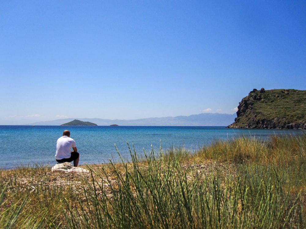 Hombre con camisa blanca sentado en la hierba verde cerca del cuerpo de agua durante el día