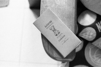 A black and white image featuring a neatly designed coffee package resting on a wooden surface. The text on the package reads 'Good Morning Starts Coffee' and includes an illustration of a coffee maker. Next to the package, part of a patterned fabric can be seen.