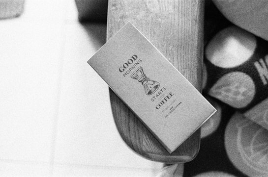 A black and white image featuring a neatly designed coffee package resting on a wooden surface. The text on the package reads 'Good Morning Starts Coffee' and includes an illustration of a coffee maker. Next to the package, part of a patterned fabric can be seen.
