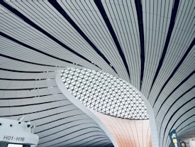 Curved architectural structure with ribbed design lines leading towards a large, circular, geometric skylight. The lower portion features a section of wall with gates marked H01-H16 and some signage.