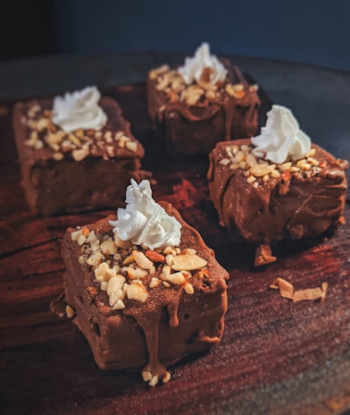 Four chocolate-covered desserts are topped with crushed nuts and a dollop of whipped cream, arranged on a dark wooden surface.