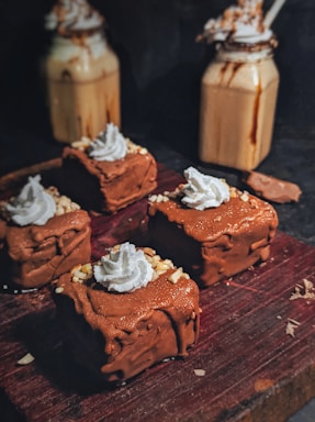 Four chocolate cakes topped with whipped cream and sprinkled with chopped nuts are arranged on a dark wooden board. In the background, there are two jars filled with creamy beverages, topped with whipped cream and drizzled with chocolate.