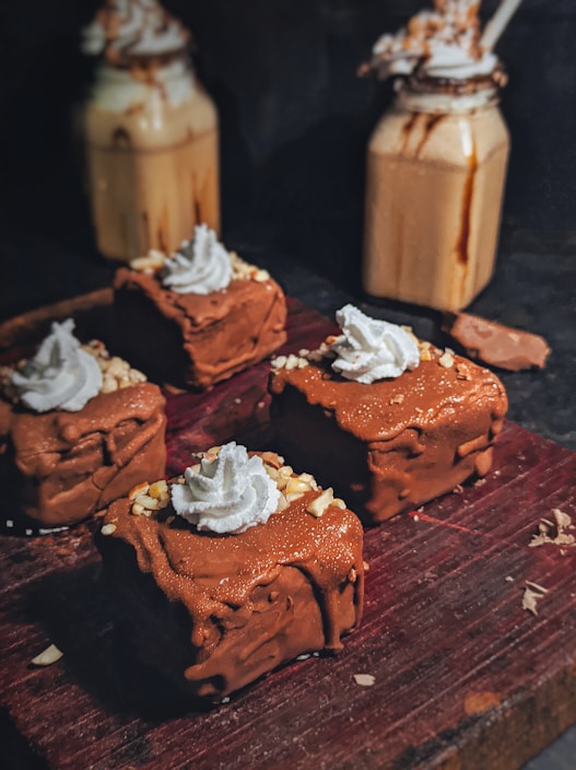 Four chocolate cakes topped with whipped cream and sprinkled with chopped nuts are arranged on a dark wooden board. In the background, there are two jars filled with creamy beverages, topped with whipped cream and drizzled with chocolate.