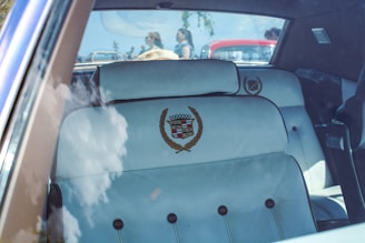 The interior of a vintage car with white leather seats, featuring embroidered crests. Reflections of clouds are visible on the windshield, and other cars and people are outside, slightly out of focus.