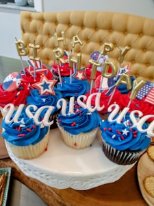 A collection of cupcakes with vibrant blue and red icing, topped with sprinkles and decorative elements. The cupcakes spell out 'HAPPY BIRTHDAY' in golden candle letters and 'USA' in white letters. Additional decorations include star-shaped picks and flags with red, white, and blue patterns. The cupcakes are arranged on a white cake stand.