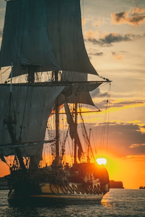 A large sailing ship is silhouetted against a vibrant sunset on the horizon. The sun casts a warm golden glow over the sky and reflects on the calm sea, highlighting the ship's tall masts and billowing sails.