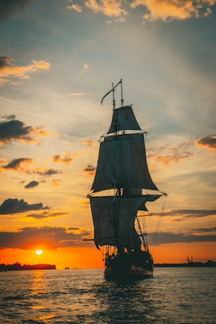 A dramatic seascape featuring a tall ship silhouetted against a fiery sunset sky.