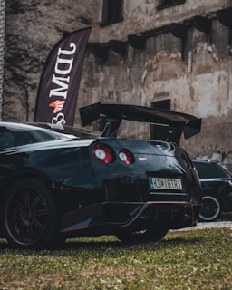 A sleek black sports car with red accents is parked on grass in front of an old, weathered stone building. The car has a large rear spoiler and distinctive round taillights. A vertical flag with the letters 'DMW' is positioned near the car.