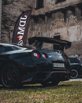 A sleek black sports car with red accents is parked on grass in front of an old, weathered stone building. The car has a large rear spoiler and distinctive round taillights. A vertical flag with the letters 'DMW' is positioned near the car.