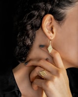 A close-up of a person's neck, ear, and hand. The person is wearing gold jewelry, including an intricate earring and matching ring, both with a similar geometric, bead-like design. The hair is styled in braids, and the person is wearing dark nail polish.