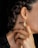 A close-up of a person's neck, ear, and hand. The person is wearing gold jewelry, including an intricate earring and matching ring, both with a similar geometric, bead-like design. The hair is styled in braids, and the person is wearing dark nail polish.